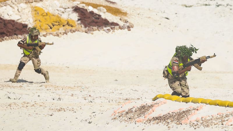 Egyptian soldiers in desert camouflage and hi-vis vests advance with rifles on a sandy training range