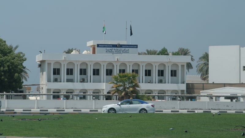 White Al Heera Police Station with a Sharjah Police G.H.Q. sign and UAE flag on the roof, a taxi passing in front