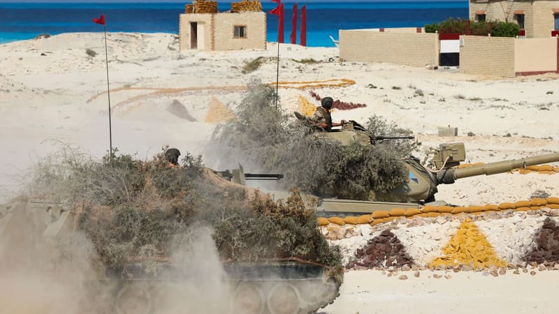 Brush-camouflaged Egyptian Army tank with a crewman in the hatch, dust rising, an Egyptian flag painted on a wall behind