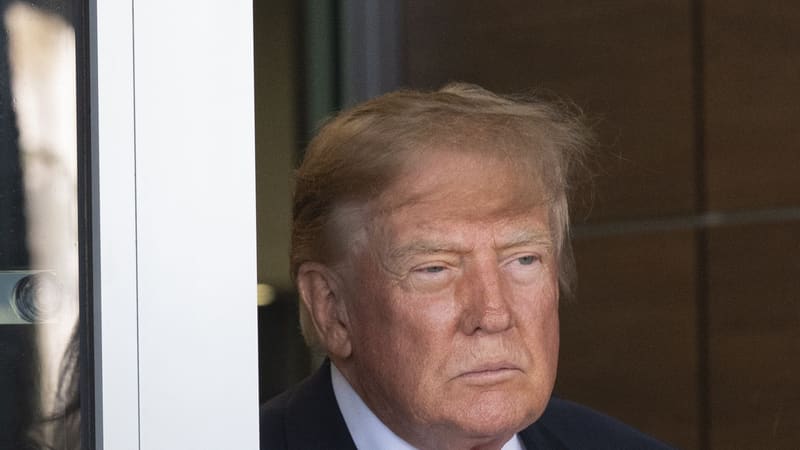Donald Trump in a glass doorway in a dark suit, looking to the side